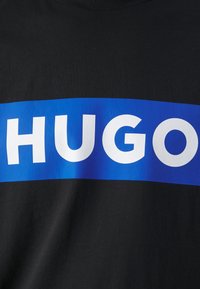 Zwarte t-shirt met een grote horizontale blauwe streep waarop het woord "HUGO" in vetgedrukte witte hoofdletters staat. Glad katoenmateriaal.