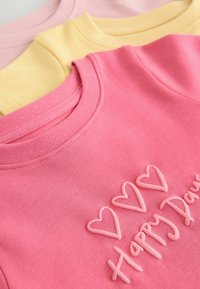Trois t-shirts à col rond en rose, jaune et rose clair. Le t-shirt rose présente des cœurs brodés et le texte "Jours Heureux". Tissu doux.