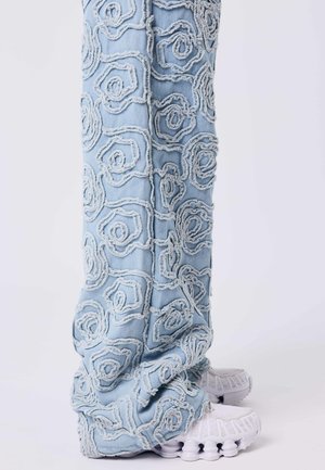 Jean bleu clair avec des motifs en spirale surélevés et effilochés, associés à des baskets blanches sur fond blanc.