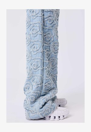 Jean bleu clair avec des motifs en spirale surélevés et effilochés, associés à des baskets blanches sur fond blanc.
