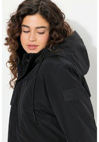 Zwarte geïsoleerde jas met een capuchon, voorzien van een ritszak en een textuurpatchlabel op de mouw. Glad materiaal en een relaxte pasvorm.