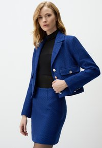 Blauer Textil-Blazer mit taillierter Passform, ausgestattet mit zwei vorderen Taschen und silbernen Knöpfen, kombiniert mit einem passenden Rock.