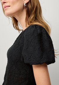 Schwarzes Oberteil aus strukturiertem Spitzenstoff, mit gepufften kurzen Ärmeln und aufwendigen floralen Mustern. Glatter Halsausschnitt und leicht taillierte Form.