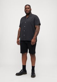 Camicia nera a maniche corte con bottoni, abbinata a pantaloni corti neri. La camicia presenta un colletto rilassato e bottoni frontali, con una texture liscia. Le scarpe nere e i calzini completano il look.