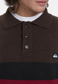 Braunes Poloshirt mit Kragen, das über einen strukturierten Strickstoff verfügt, drei Knöpfe hat und horizontale Streifen in Schwarz und Rot mit einem Logopatch an der Seite aufweist.