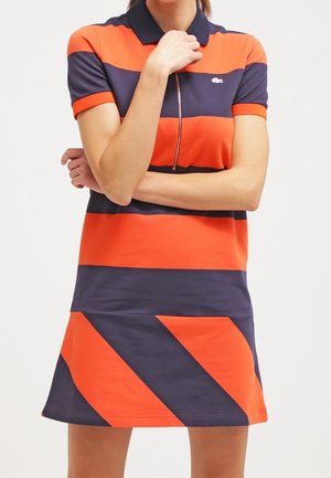 Kobieta w sukience polo z krótkim rękawem, zdobionej pomarańczowo-granatowymi poziomymi paskami i zamkiem z przodu, z założonymi ramionami i ręką przy brodzie.