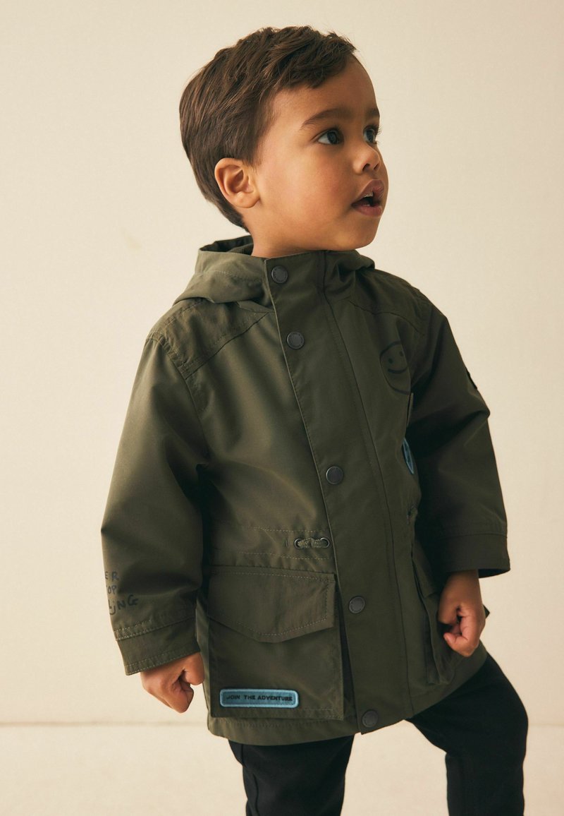 Chaqueta impermeable verde para niños con capucha, botones a presión, dos bolsillos delanteros y parche azul con la frase "Únete a la aventura".