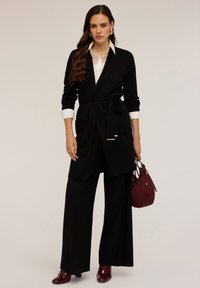 Gilet ceinturé noir sur chemise blanche, associé à un pantalon large noir et un sac à main bordeaux, agrémenté de bottines à talons bordeaux brillantes.