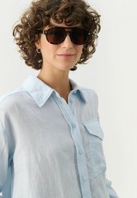 Black tortoiseshell sunglasses with gradient lenses. Light blue linen shirt with a collar and a front pocket, featuring a relaxed fit and texture.