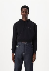 Svart cropped hettegenser med snøring, med en hvit "SALOMON" logo, kombinert med høy midje mørk denimjeans med grønne detaljer.