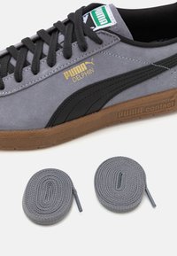 Puma Delphin sneakers in grey suede with black accents, featuring a gold logo and brown rubber sole. Includes two grey shoelaces.