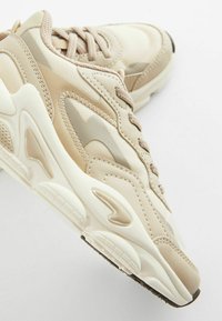 Zapatillas deportivas beige que combinan materiales texturizados y lisos, con costuras visibles y una gruesa suela de goma con un diseño de trazado.