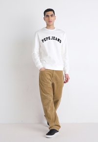 Witte sweatshirt met zwarte letters "PEPE JEANS", gecombineerd met tan corduroy broek en zwarte sneakers, op een effen achtergrond.