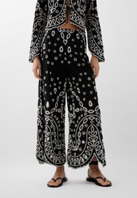 Zwarte wijde broek met witte bloemborduursels en uitsnijdingen, gecombineerd met een bijpassende cropped jas en zwarte sandalen.