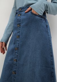 Gonna di denim con chiusura a bottoni frontale, tasche laterali e vita aderente. Il tessuto è di un blu medio con una texture liscia.