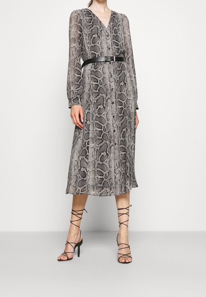 Femme portant une robe noire et grise à manches longues, boutonnée, imprimé serpent, avec une ceinture noire et des sandales à talons hauts noires à lacets.
