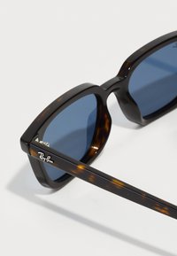 Lunettes de soleil en écaille de tortue avec des verres bleu foncé, une monture épaisse et un détail de logo argenté sur la branche. Texture lisse, forme rectangulaire audacieuse.