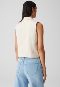 Top corto senza maniche in tessuto beige chiaro, con colletto e una texture liscia, abbinato a jeans azzurri a vita alta.