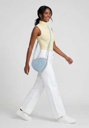 Woman walking with white pants, sleeveless yellow turtleneck top, white sneakers, and a light blue heart-shaped crossbody bag.