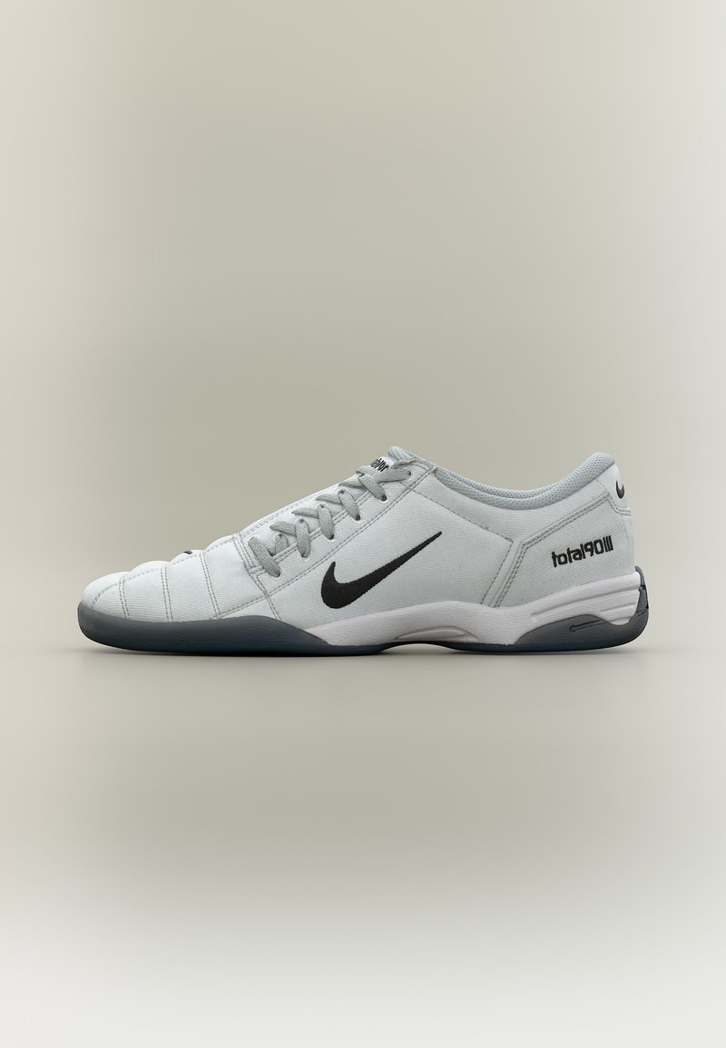 Chaussure de football basse Nike blanche et grise avec logo swoosh noir, lacets et texte « total90 III » sur le talon, vue de profil.