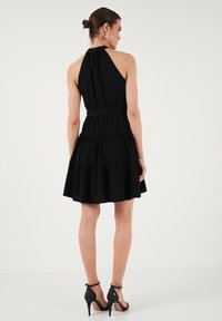 Robe noire à bretelles avec un corsage ajusté, dos froncé, jupe à volants et texture subtile ; associée à des sandales à talons hauts noires.