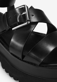 Sandalo platform nero con fasce in pelle lucida, una fibbia metallica e una suola testurizzata. Il design enfatizza linee geometriche audaci.