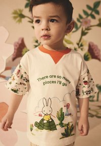 Balti marškinėliai su oranžiniu V formos iškirpte ir spalvotomis trumpomis rankovėmis. Dizainas vaizduoja animacinį kiškį ant vėžlio, tekstas: "Yra tiek daug vietų, kurias aplankysiu."