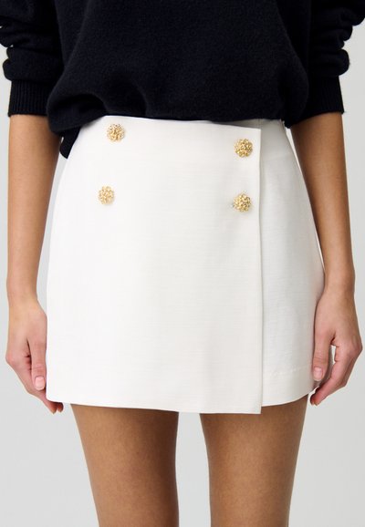 Minifalda blanca con un diseño estructurado, que presenta cuatro botones decorativos en forma de flor en tono dorado y una tela de textura suave.