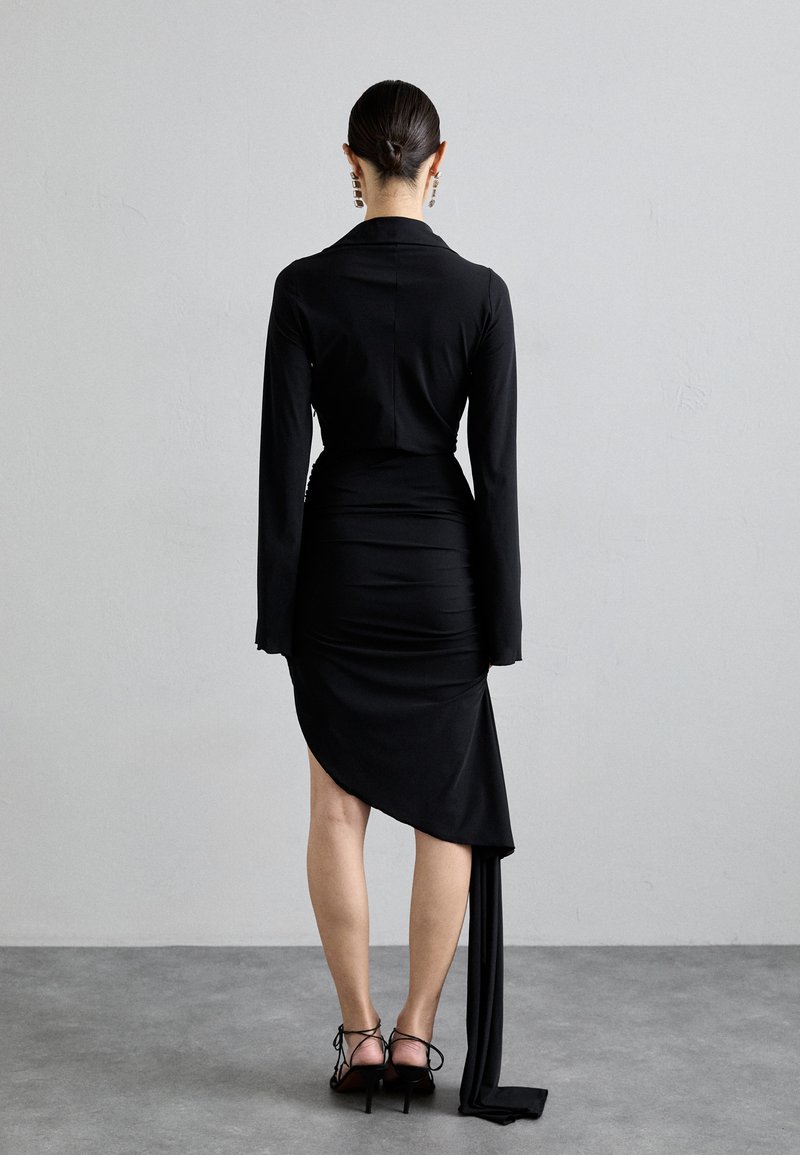 Zwarte jurk met lange mouwen, aansluitende lijfje en asymmetrische zoom. Bevat gedrapeerde stof en een vloeiende staart. Paar zwarte strappy hakken.