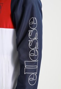 Marinblå jacka med en vit logotyp på ärmen och ett röd och vit färgblock design på den övre delen. Slät tygtextur.