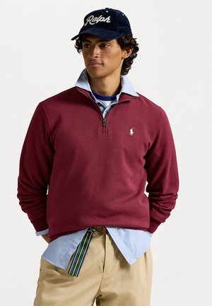 Burgunderfarget hettegenser med glidelås og en liten logo, lagd over en lyseblå skjorte med krage. Mørk marineblå caps med "Ralph"-tekst. Elfenbensfargede bukser.