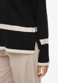 Melnas krāsas džemperis ar platām krēmkrāsas horizontālām svītrām, garām piedurknēm, sānu šķēlumiem un mīkstu tekstūru, kombinēts ar gaiši bēšām biksēm.