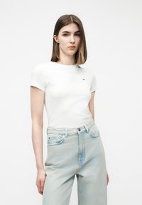 Νεαρή γυναίκα με ίσια καστανά μαλλιά φοράει εφαρμοστό λευκό κοντομάνικο μπλουζάκι και ανοιχτό μπλε τζιν με ψηλή μέση, στέκεται μπροστά σε λευκό φόντο.