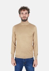 Maglione beige dolcevita in tessuto leggero a coste. Presenta maniche lunghe e una vestibilità aderente, abbinato a jeans blu scuro.