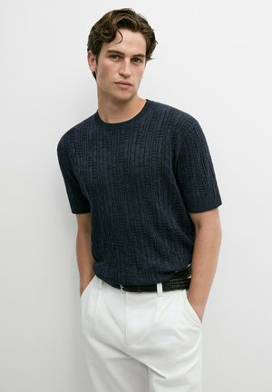 Giovane uomo con capelli castani mossi indossa un maglione scuro a maniche corte con motivo, infilato in pantaloni bianchi, con le mani nelle tasche.