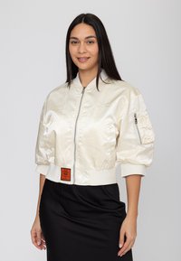 Donna che indossa una giacca bomber corta in raso color crema con tasca con zip sulla manica e una gonna nera, che sorride leggermente, su uno sfondo semplice.