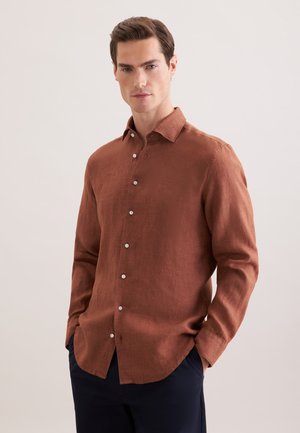 Mann trägt ein rostfarbenes, langärmliges Hemd mit Kragen und Knöpfen, Hände in den Taschen, steht vor einem schlichten hellen Hintergrund.