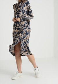 Marinblå klänning med blommönster i beige, långa ärmar, hög slits och knäppning framtill. Matchad med vita sneakers för kontrast.