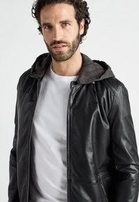 Svart läderjacka med grå huva, slät textur, dragkedja, sidfickor och snörning i huvan. Bärs över en vit t-shirt.