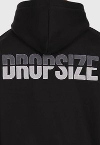 Haut de survêtement noir avec de grandes lettres texturées "DROPSIZE" brodées sur le haut du dos, divisé horizontalement en deux teintes contrastées.