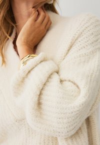 Cremefarvet strikket sweater med et struktureret mønster, der har rullede manchetter og en dyb V-udskæring. Et chunky guldbelagt armbånd er synligt på håndleddet.