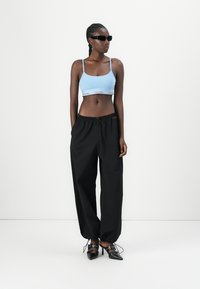 Lichtblauwe bralette met logo-band, gecombineerd met zwarte, loszittende broeken. Model draagt een paar zwarte zonnebrillen en zwarte hakken.