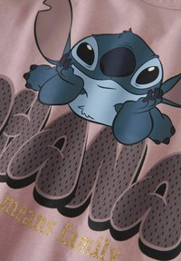 Różowy materiał z rysunkowym bohaterem o niebieskiej skórze i dużych oczach. Napis brzmi "Ohana oznacza rodzinę" w ciemnoszarym kolorze z złotymi akcentami.