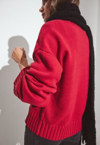 Suéter de lana rojo con dobladillo y puños acanalados, combinado con una bufanda negra. La textura parece gruesa y acogedora, destacando su diseño cálido.