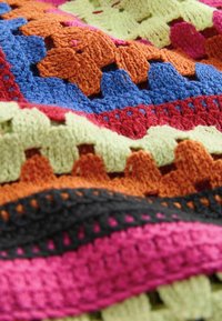 Višebojni pleteni pokrivač s istaknutom narančastom, crvenom, plavom, ružičastom i zelenom pređom u heklanoj teksturi s izraženim uzorkom.