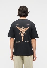 Άτομο που φοράει μαύρο T-shirt με μια μεγάλη μπεζ φιγούρα αγγέλου και κείμενο που αναφέρεται στο "In Utero" των Nirvana στην πλάτη.