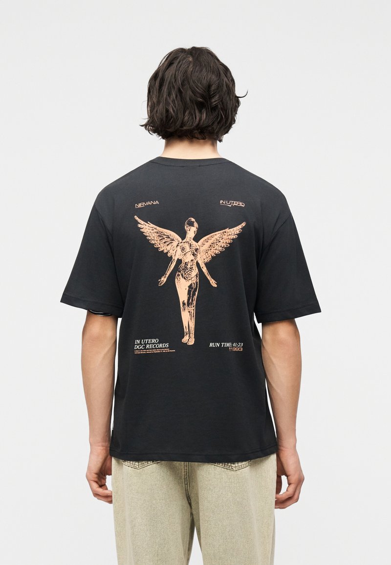 Άτομο που φοράει μαύρο T-shirt με μια μεγάλη μπεζ φιγούρα αγγέλου και κείμενο που αναφέρεται στο "In Utero" των Nirvana στην πλάτη.