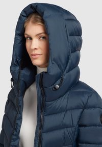 Marinblå puffer-jacka med en stor, justerbar huva, dragkedjestängning fram och quiltat mönster. Har svarta dragkedjor och runda detaljer.