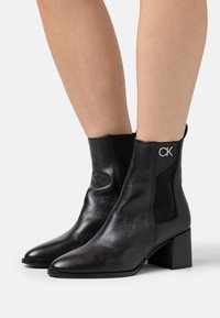 Bottines en cuir noir avec une finition texturée, un talon bloc et des panneaux latéraux élastiques. Présentent un logo CK sur le côté extérieur.