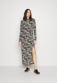 Vestido de manga longa com padrão preto e branco e fenda lateral; usado com tamancos pretos. Apresenta um colarinho alto e um tecido fluido e texturizado.